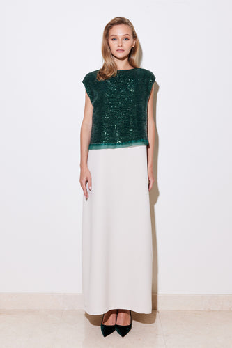 A woman standing against a white background, wearing a cream-colored heavy crepe dress with a round neck and 1/4 sleeves. The dress features a dramatic deep emerald green sequin embellished tulle fabric and green trims.