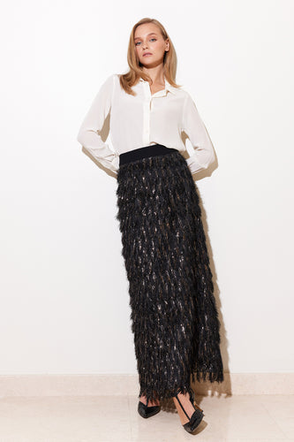 A model wearing a straight cut black skirt with shaggy metallic threads, paired with a white top. The skirt has a back slit and a black waistband with a zipper.