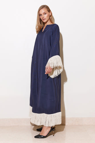 A woman standing in a navy blue dress with long sleeves adorned with cream rope tassels, posing against a white background.