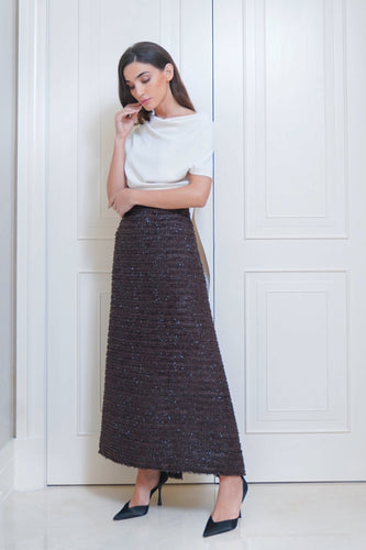 A woman standing in a doorway wearing a white top and a brown fringed skirt with striped detailing. She has her hand to her face and is wearing black shoes.