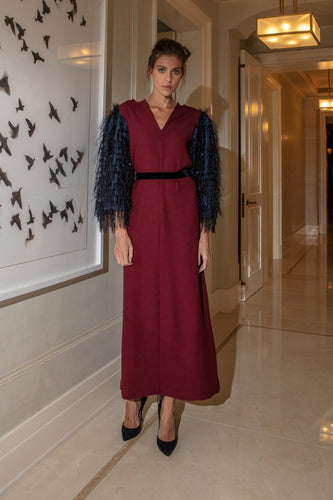 A woman standing in a maroon dress with v neckline and navy blue sleeves, featuring two side slits, and an optional thin black velvet belt.